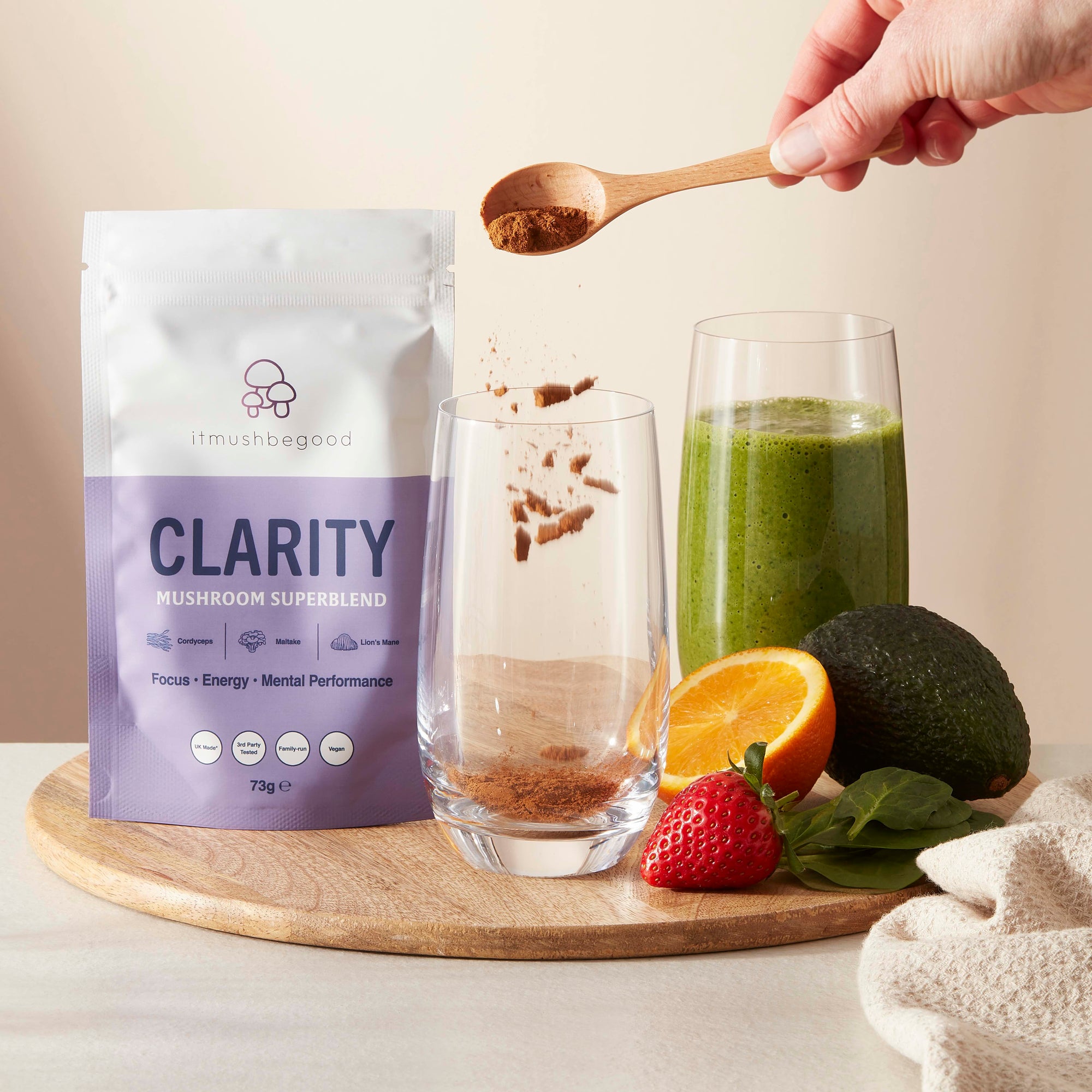 Clarity powder being spooned into glass next to a green smoothie and fruit on a chopping board